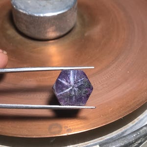 May include: A faceted, hexagonal purple gemstone with a six-pointed star pattern held by tweezers. The gem is set against a copper-colored surface with a silver metal object in the background. The gem is a deep purple color.