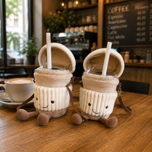 Könnte beinhalten: Zwei plüschige becherförmige Taschen in Beige und Creme mit gerippter Textur, gestickten lächelnden Gesichtern und baumelnden Beinen. Jede Tasche hat einen durchsichtigen Kunststoffdeckel, einen weißen Strohhalm und einen dünnen braunen Schulterriemen.
