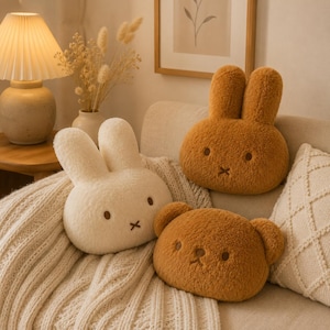 May include: Three plush cushions shaped like animal heads with soft, fuzzy textures. One cushion is cream-coloured with long rabbit ears, one is golden-brown with long rabbit ears, and one is brown with rounded bear ears, all featuring embroidered faces.