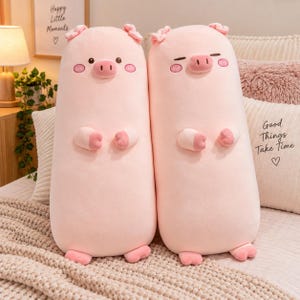 May include: Two cylindrical, light pink plush pig pillows. The left pig has open black eyes and a smiling mouth. The right pig has closed, curved eyes. Both feature small ears, a snout, two small arms, and two feet, all in a soft fabric.