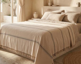 May include: A beige bedspread with dark blue stripes and fringe detailing covers a bed with matching pillows. The room features a large window with sheer curtains, a stone floor, and neutral-toned walls, creating a serene bedroom setting.