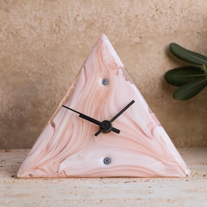 May include: A triangular clock with a pink and white swirled pattern. The clock has black hands and two small, decorative blue accents. The clock is a unique decorative item.