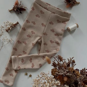 Peut inclure: Un legging beige pour bébé, avec une texture en nid d'abeille et un motif de petits cœurs bruns. Le legging est présenté sur une surface blanche, avec des fleurs séchées et du matériel de couture en arrière-plan.
