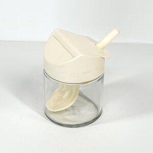 May include: A clear glass spice jar with a cream-colored lid and a small spoon inside. The lid has a sliding mechanism and a small handle. The jar is designed for easy dispensing of spices and seasonings.