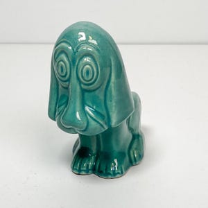 May include: A teal ceramic dog figurine with large, expressive eyes and long, floppy ears. The dog is in a seated position, with its front paws visible. The figurine has a glossy finish and is likely a decorative item.
