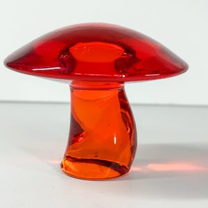 May include: A vibrant, red glass mushroom figurine. The mushroom has a rounded cap and a curved stem, with a glossy finish. The color transitions from a deep red on top to a bright orange at the base. The figurine is a decorative item.