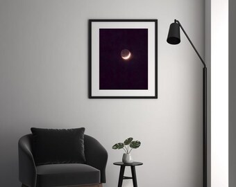 May include: A framed print featuring a crescent moon against a dark purple background. The artwork is displayed above a dark gray armchair, a small black side table with a plant, and a black floor lamp. The room has a minimalist design.