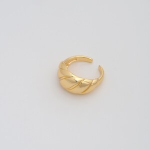 Puede incluir: Un ear cuff dorado con un diseño texturizado y acanalado. El brazalete abierto se curva alrededor de la oreja, ofreciendo un accesorio moderno y elegante. La joya se coloca sobre un fondo blanco liso, resaltando su elegante forma.