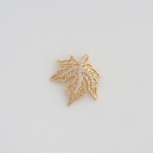May include: A gold-colored maple leaf pendant with a delicate, openwork design. The pendant is adorned with small, clear crystals that form a cross-like pattern across the leaf. The jewelry piece is set against a plain white background.