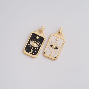 May include: Two rectangular pendants with gold-colored frames. One pendant has a black background with a white eye, moon, and stars. The other pendant has a white background with a black eye, moon, and stars. Both pendants have a gold-colored loop at the top.