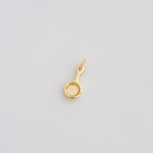 May include: A small, gold-colored charm in the shape of a frying pan. The charm has a circular pan with a handle and a small loop at the top for attaching to a chain or bracelet. The charm is made of a shiny, reflective metal.