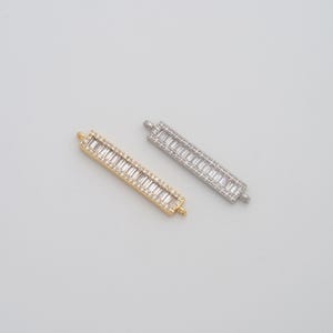 May include: Two rectangular jewelry components, one gold-toned and one silver-toned, each encrusted with small, clear, rectangular-cut stones. The components have small loops at each end for attachment. The background is a plain, light gray.