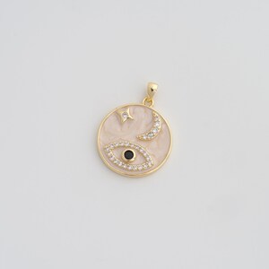 May include: Gold-toned pendant with a circular design featuring an eye, crescent moon, and star, all adorned with clear crystals. The pendant has a light pink and white marbled background.