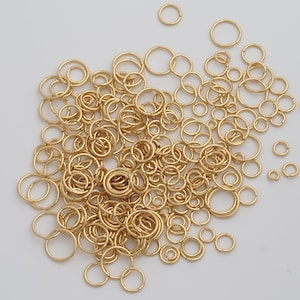 May include: A collection of gold-colored jump rings of various sizes, scattered across a white surface. The rings are circular, with a split for attaching to other jewelry components. The image showcases a variety of sizes, from small to medium.