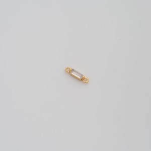 May include: A small, gold-colored jewelry component with a rectangular, clear crystal center. The component has small loops on each end for attachment. The background is a plain, light-colored surface.