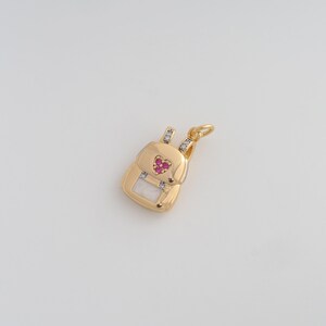 May include: A gold-colored backpack charm with a heart-shaped design in the center, adorned with small red gemstones. The charm has a rectangular shape with a white rectangular accent at the bottom. Small clear gemstones are set on the top and sides.