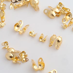 May include: A collection of shiny gold-colored jewelry findings, including crimp end caps and other small components. The pieces are scattered across a white surface, showcasing their reflective quality and intricate designs. These findings are likely used for jewelry making.