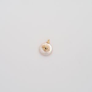 May include: A gold-colored pendant with a white, round pearl base. The pendant features a gold-colored eye design with a black center and surrounding clear crystals. The pendant has a small gold loop at the top for a chain.
