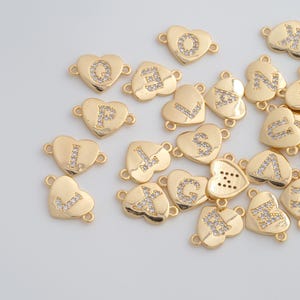 May include: Gold-coloured heart-shaped charms with letters of the alphabet, each encrusted with small, clear crystals. The charms have loops on each side for attaching to jewellery. The letters are in a serif font.