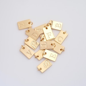 May include: Gold-colored rectangular charms with embossed letters of the alphabet. Each charm has a small loop at the top for attaching to a chain or bracelet. The charms are scattered on a white surface.