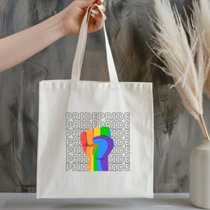 Könnte beinhalten: Eine weiße Canvas-Tragetasche mit einem Regenbogen-Faust-Motiv und dem Wort "PRIDE", das mehrmals in schwarzer Schrift wiederholt wird.
