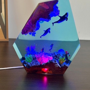 May include: A decorative resin lamp with an underwater scene. The lamp features a blue and purple design with orcas, divers, and coral. The lamp has a geometric shape and emits a soft, colorful glow.