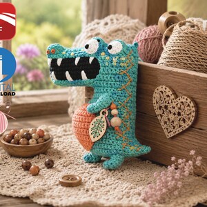 May include: A turquoise and orange crocheted crocodile toy with large white eyes and a black mouth featuring white teeth. The crocodile holds a wooden leaf-shaped charm and wooden beads, with orange stitching along its back and side.
