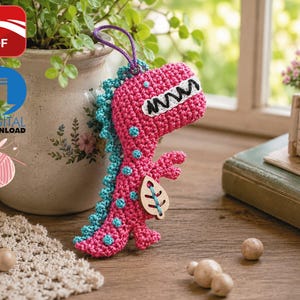 Peut inclure: Un dinosaure au crochet rose vif avec un dos et des taches bleu sarcelle. Le dinosaure présente une bouche blanche avec des dents noires brodées et une petite breloque en bois en forme de feuille attachée à la poitrine avec une boucle violette.