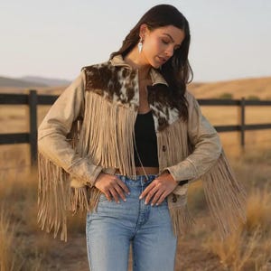 Peut inclure: Veste en daim beige avec longues franges et empiècement en peau de vache. La veste a une fermeture boutonnée. Le fond présente une clôture en bois et un champ. La veste est portée avec un jean bleu.