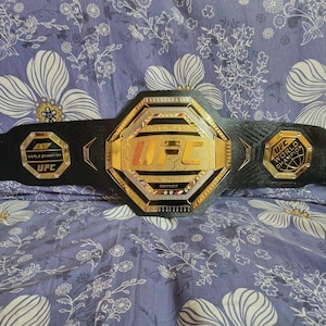 May include: A black leather UFC World Championship belt with gold accents. The belt features a large octagon-shaped buckle with the UFC logo and the words "World Champion" in gold lettering.