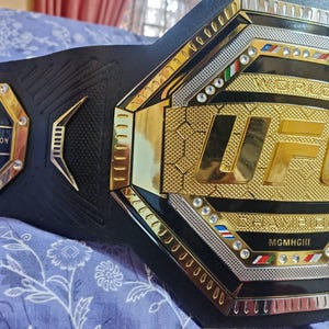 May include: A black and gold UFC World Champion belt with a large octagon-shaped center plate. The center plate features the UFC logo in gold with the words "World Champion" and "MGM Grand" in smaller gold lettering. The belt is adorned with rhinestones and has a black leather strap.