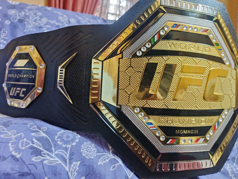 May include: A black and gold UFC World Champion belt with a large octagon-shaped center plate. The center plate features the UFC logo in gold with the words "World Champion" and "MGM Grand" in smaller gold lettering. The belt is adorned with rhinestones and has a black leather strap.