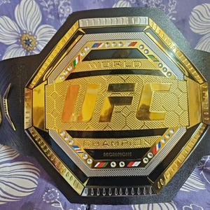 May include: A black leather UFC championship belt with a gold octagon-shaped plate featuring the UFC logo and the words "World Champion".