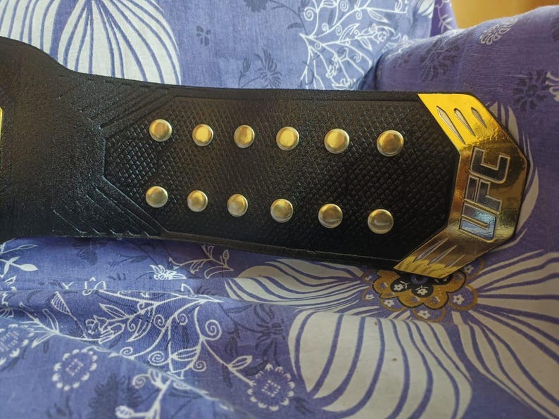 May include: A black leather belt with gold accents and silver studs. The belt has the word "UFC" in gold lettering on the side.