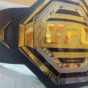 May include: A black and gold UFC championship belt with the words "World Champion" and "MCMXCIII" on it.