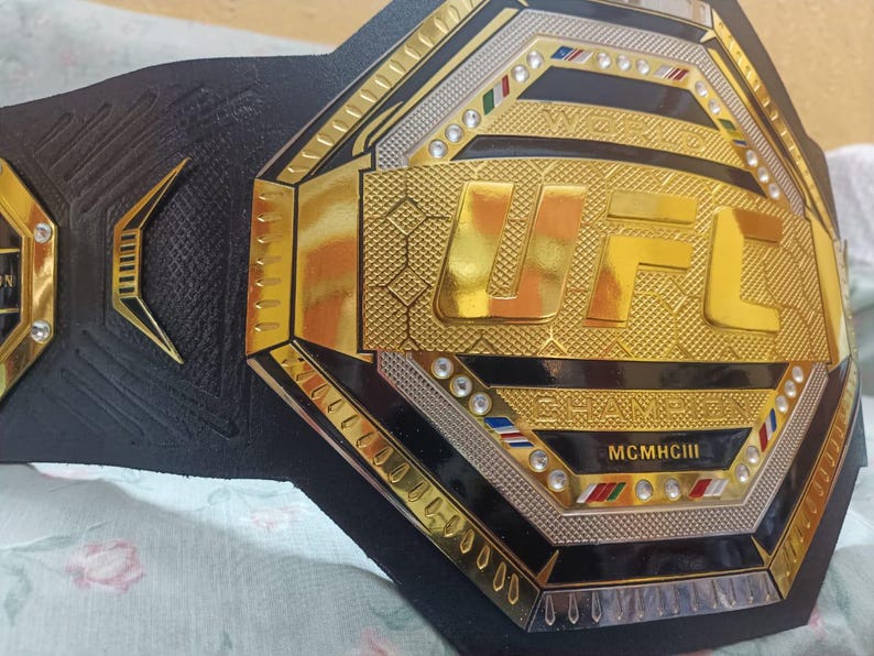 May include: A black and gold UFC championship belt with the words "World Champion" and "MCMXCIII" on it.