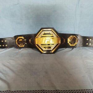 May include: A black UFC championship belt with gold accents. The belt features the UFC logo and the words "World Heavyweight Champion".