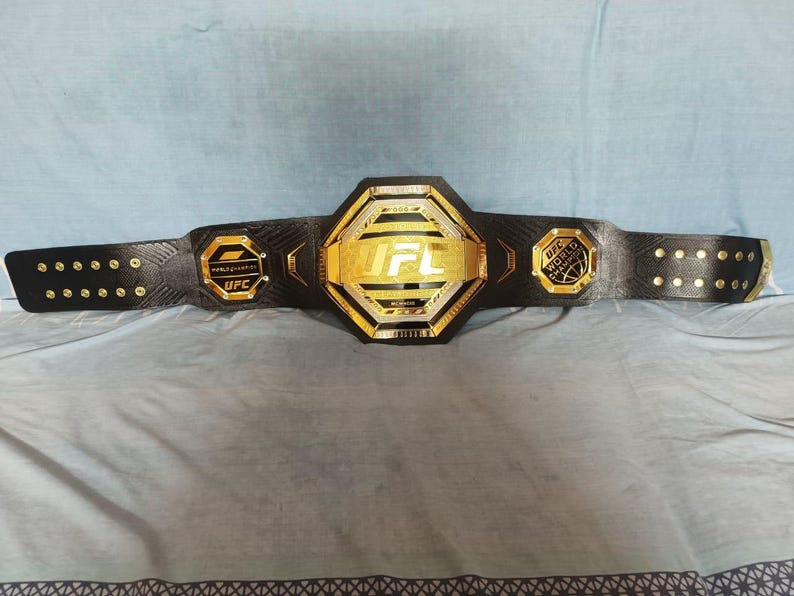 May include: A black UFC championship belt with gold accents. The belt features the UFC logo and the words "World Heavyweight Champion".