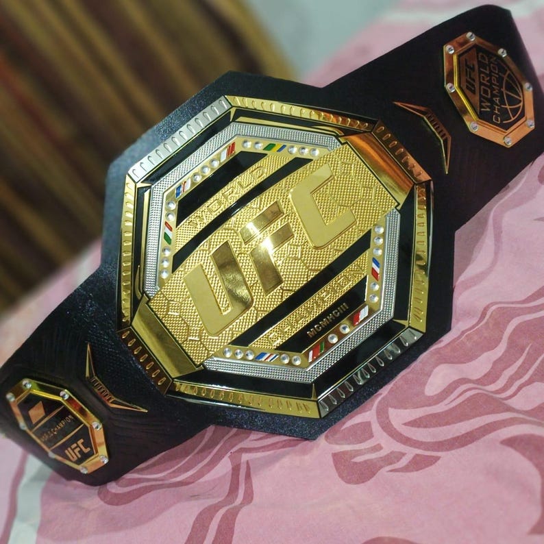 May include: A gold and black UFC World Champion belt with a black leather strap and a large octagon-shaped buckle with the UFC logo and the words "World Champion" in gold lettering.