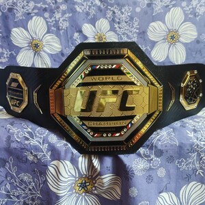 May include: A black UFC World Champion belt with a gold octagon-shaped buckle. The buckle features the UFC logo and the words "World Champion".