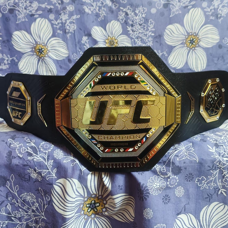 May include: A black UFC World Champion belt with a gold octagon-shaped buckle. The buckle features the UFC logo and the words "World Champion".