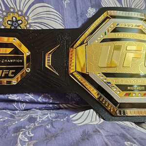 May include: A gold and black UFC World Champion belt with the words "World Champion" and "UFC" on the front. The belt has a black leather strap and a gold buckle with the UFC logo.