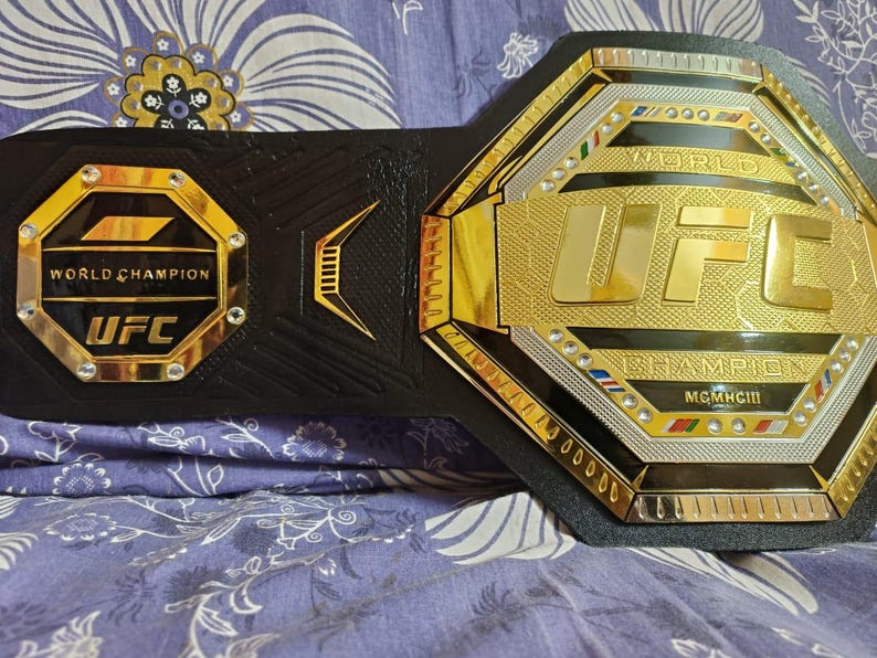 May include: A gold and black UFC World Champion belt with the words "World Champion" and "UFC" on the front. The belt has a black leather strap and a gold buckle with the UFC logo.