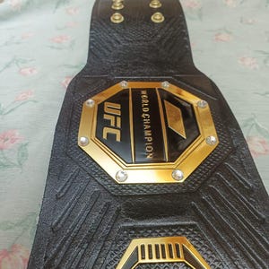 May include: A black leather UFC championship belt with gold accents and rhinestones. The belt features the UFC logo and the words "World Champion".
