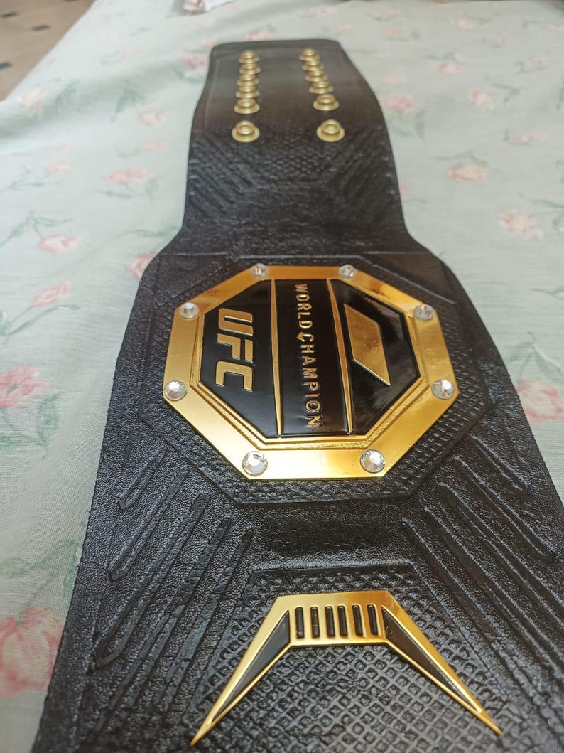 May include: A black leather UFC championship belt with gold accents and rhinestones. The belt features the UFC logo and the words "World Champion".