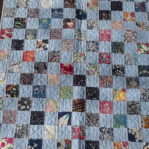 Può includere: Una trapunta patchwork con sfondo azzurro e una griglia di quadrati colorati. I quadrati presentano tessuti floreali e fantasia in vari colori, tra cui rosa, blu e verde. La trapunta misura circa 91 cm di larghezza e 122 cm di lunghezza.