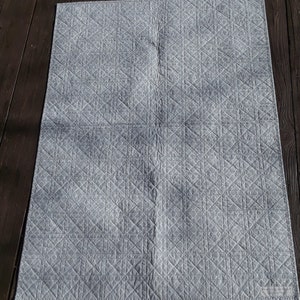 Pu&ograve; includere: Una coperta trapuntata grigio chiaro con un motivo geometrico. La coperta &egrave; rettangolare e presenta un motivo a rombi cuciti sulla sua superficie. Il tessuto appare morbido e le cuciture sono dettagliate. La coperta &egrave; appoggiata su una superficie di legno scuro.