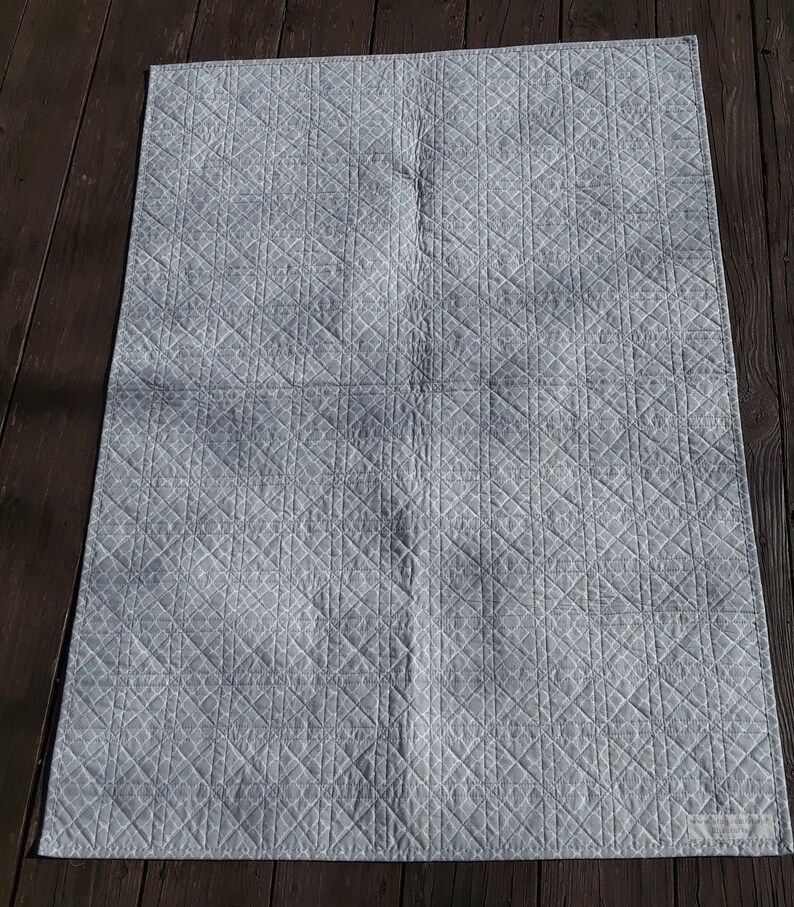 Pu&ograve; includere: Una coperta trapuntata grigio chiaro con un motivo geometrico. La coperta &egrave; rettangolare e presenta un motivo a rombi cuciti sulla sua superficie. Il tessuto appare morbido e le cuciture sono dettagliate. La coperta &egrave; appoggiata su una superficie di legno scuro.