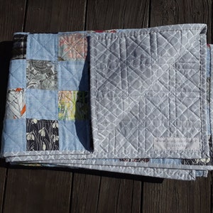 Pu&ograve; includere: Una trapunta fatta a mano con un design patchwork. La trapunta presenta quadrati di tessuto azzurro e quadrati di tessuto fantasia in vari colori. Il retro &egrave; grigio tinta unita con un motivo geometrico bianco. La trapunta &egrave; piegata.