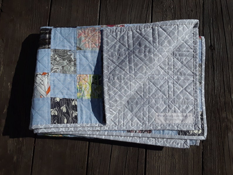 Pu&ograve; includere: Una trapunta fatta a mano con un design patchwork. La trapunta presenta quadrati di tessuto azzurro e quadrati di tessuto fantasia in vari colori. Il retro &egrave; grigio tinta unita con un motivo geometrico bianco. La trapunta &egrave; piegata.